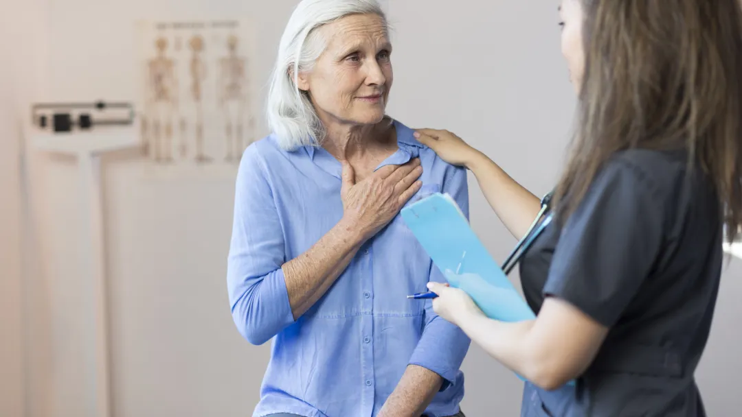 A senior woman clutching her chest, with a tech taking her blood pressure.