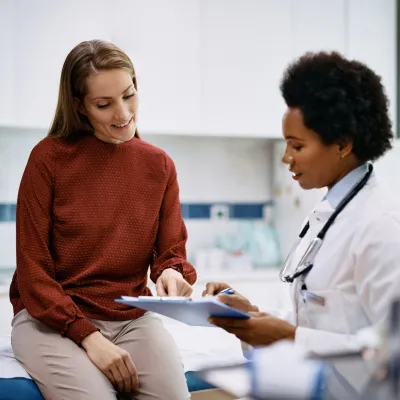 A Doctor Speaks to Her Patient in an Exam Room While Going Over Her Charts on a Laptop.