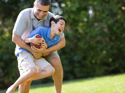 A father playfully tackles his son as they play football outside.
